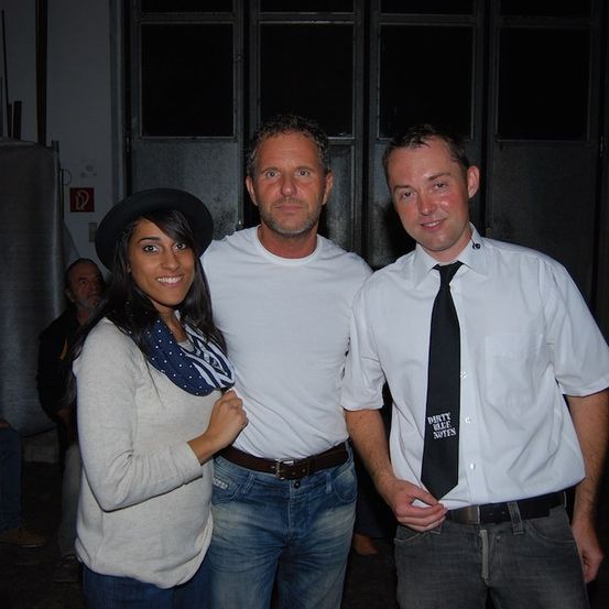 Drei Menschen stehen eng beieinander in einem Raum und lächeln für ein Foto. Eine Frau mit Schal und Hut, ein Mann in einem weißen T-Shirt und Jeans und ein weiterer Mann in einem weißen Hemd mit schwarzer Krawatte.