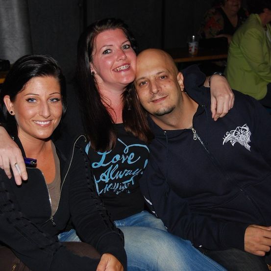 Drei Menschen sitzen zusammen und lächeln für ein Foto. Die Frau links hat einen Ring am Finger, die mittlere Frau hat Zahnspangen und der Mann rechts hat ein weißes Logo auf seiner Jacke.