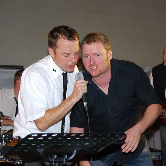 Zwei Männer treten auf der Bühne auf, einer singt in ein Mikrofon, während der andere zusieht. Dahinter spielen ein Schlagzeuger und ein Gitarrist.