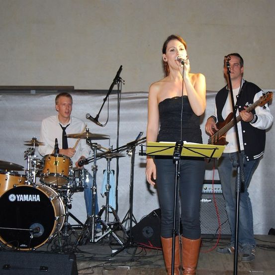 Eine Frau singt in ein Mikrofon auf der Bühne, begleitet von einem Mann, der Gitarre spielt, und einem anderen, der Schlagzeug spielt.