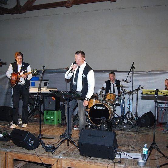 Eine Band spielt auf einer hölzernen Bühne mit einem Yamaha-Schlagzeug, einer Tastatur und Gitarren. Der Leadsänger singt in ein Mikrofon.