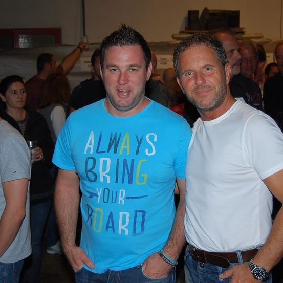 Zwei Männer stehen bei einer gesellschaftlichen Veranstaltung nebeneinander, einer trägt ein blaues T-Shirt mit dem Text 'Always Bring Your Board' und der andere ein weißes T-Shirt. Sie sind von einer Menge Menschen umgeben.