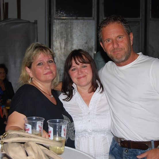 Drei Menschen lächeln und posieren für ein Foto. Der Mann trägt ein weißes T-Shirt und Jeans. Die Frau auf der linken Seite trägt ein schwarzes T-Shirt und hält einen Becher. Die Frau in der Mitte trägt eine weiße Bluse.