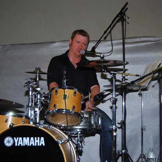 Ein Mann spielt Schlagzeug auf der Bühne mit einem Yamaha-Schlagzeug, trägt ein schwarzes Hemd und Jeans, mit Mikrofonen und Becken um ihn herum.