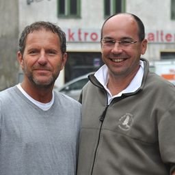 Zwei Männer lächeln und posieren für ein Foto vor einem Gebäude mit einem Schild, das 'ktro alters' steht.