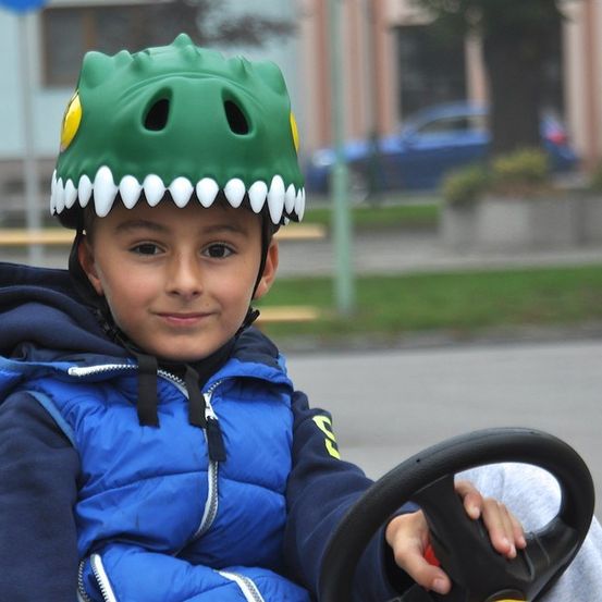 Ein junger Junge mit einem blauen Jackett und einem grünen Dinosaurierhelm sitzt in einem Spielzeugauto, lächelt und hält das Lenkrad.