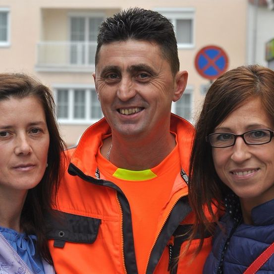 Drei Menschen stehen eng beieinander draußen. Ein Mann in einer orangefarbenen Weste lächelt in die Kamera. Die beiden Frauen neben ihm schauen geradeaus.