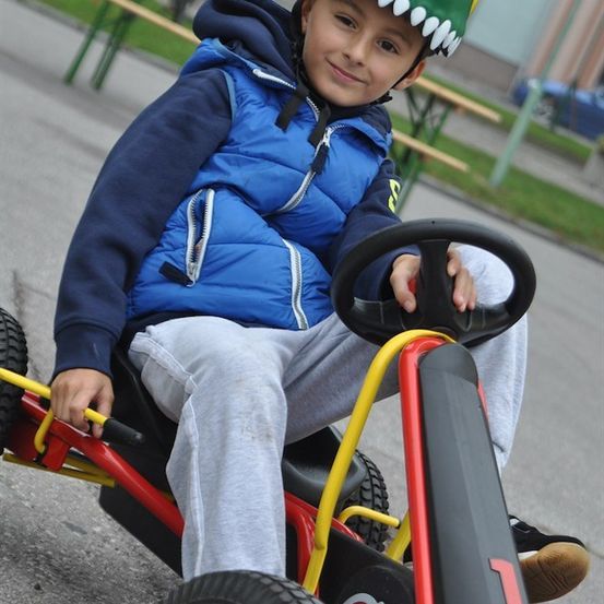 Ein junger Junge in einem blauen Jacken und einem Dinosaurier-Helm sitzt in einem roten Go-Kart, lächelt und hält das Lenkrad.
