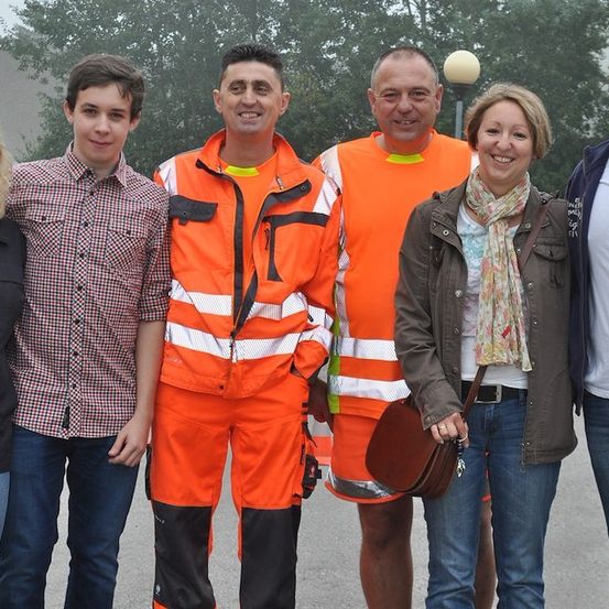 Eine Gruppe von Menschen, darunter zwei Männer in orangefarbenen Sicherheitswesten, steht draußen zusammen. Eine Frau in einer braunen Jacke hält eine braune Handtasche. Im Hintergrund befinden sich Bäume und ein Gebäude.