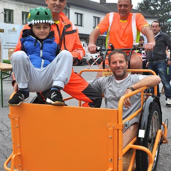 Drei Männer in orangefarbenen Westen und ein junger Junge in einer blauen Jacke und einem grünen Helm fahren ein Dreirad. Der Junge sitzt vorne, während die Männer hinter ihm sitzen.
