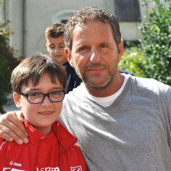 Ein Mann mit Bart und ein Junge in einem roten Jackett mit Brille stehen eng beieinander und lächeln für ein Foto. Hinter ihnen steht ein Junge in einem blauen Shirt in der Nähe einiger Pflanzen.