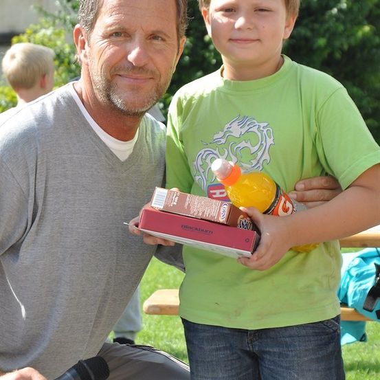 Ein Mann und ein Junge posieren für ein Foto. Der Junge hält eine Schachtel und eine Flasche. Der Mann hält ein Mikrofon.