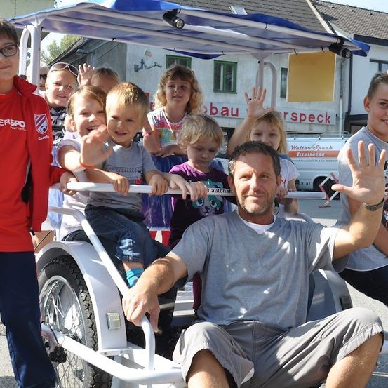 Ein Mann sitzt auf einem Dreirad mit Kindern um ihn herum, winkt und lächelt, vor einem Gebäude mit einem Schild, das 'Walter Speck' steht.