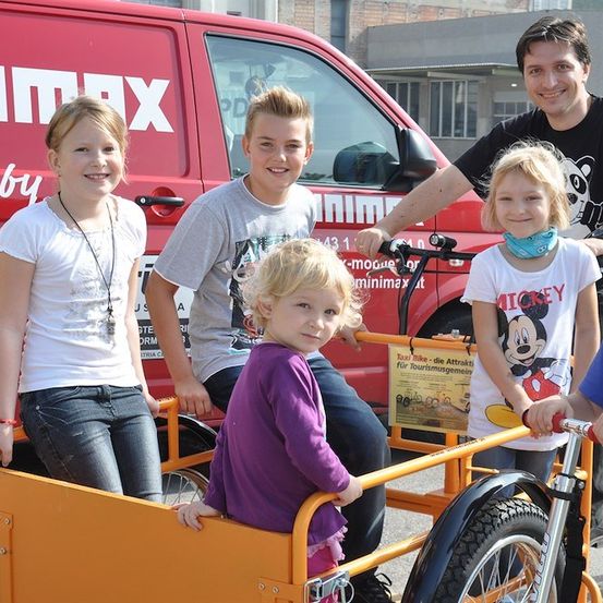 Eine Gruppe von Kindern und zwei Erwachsenen stehen neben einem roten Van. Die Kinder sitzen auf einem gelben Fahrrad mit einem Korb vorne. Der Mann auf der rechten Seite hält das Lenkrad des Fahrrads. Die Kinder lächeln und posieren für ein Foto. Der Van hat den Text 'NIMMAX' darauf.