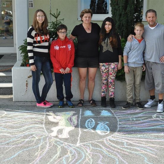 Eine Familie mit fünf Personen steht vor einem Gebäude mit einer Kreidezeichnung auf dem Boden. Die Familie besteht aus zwei Erwachsenen und drei Kindern. Die Erwachsenen tragen Freizeitkleidung. Die Kinder tragen Turnschuhe.
