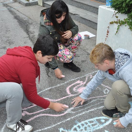 Drei Kinder zeichnen mit Kreide auf dem Boden, eines hält eine blaue Kreide. Das Mädchen in der Mitte sitzt und schaut auf die Zeichnung. Der Junge rechts kniet und zeigt auf die Zeichnung. Der Junge links hockt und hält eine rosa Kreide.