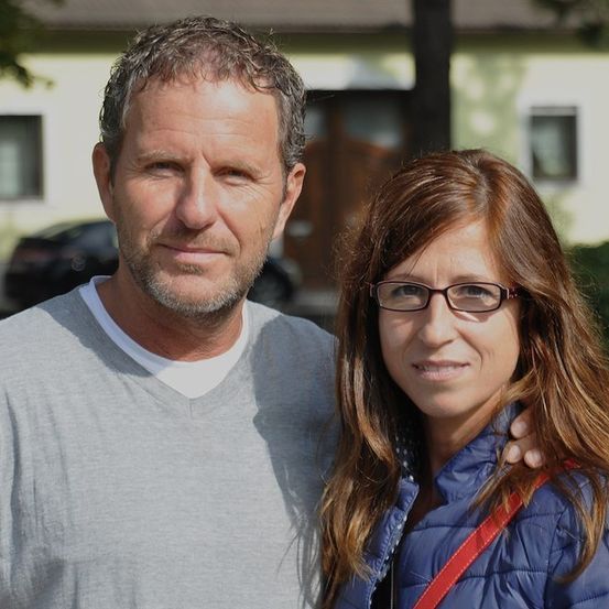 Ein Mann mit Bart und eine Frau mit Brille stehen draußen und lächeln für ein Foto. Hinter ihnen steht ein Haus mit einem Auto davor.