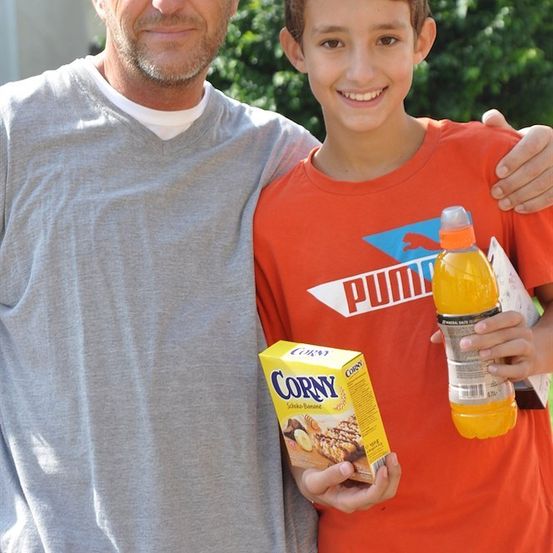 Ein Mann und ein Junge stehen lächelnd zusammen, der Junge hält eine Flasche Orangensaft und eine Schachtel Corny-Müsli.