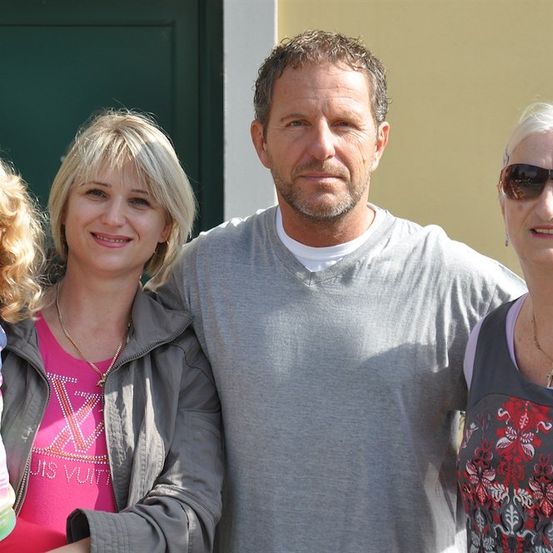 Eine vierköpfige Familie steht gemeinsam vor einer grünen Tür. Der Mann hat einen Bart und trägt ein graues T-Shirt. Die Frau links hat blonde Haare und trägt ein pinkes Shirt. Die Frau rechts trägt eine Sonnenbrille und ein Blumenshirt. Das Kind trägt ein pinkes Shirt mit Herzen.