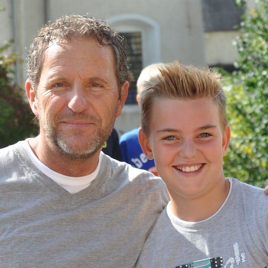 Ein Mann und ein Junge stehen zusammen draußen und lächeln für ein Foto. Der Mann hat einen Bart und trägt ein graues Shirt, während der Junge kurze Haare und ein graues T-Shirt trägt. Hinter ihnen ist ein Gebäude mit einem Fenster und etwas Grün.
