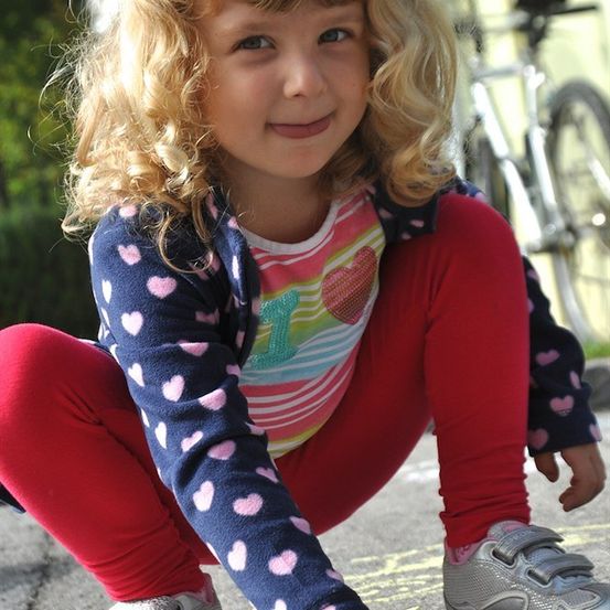 Ein kleines Mädchen mit blonden Locken kniet auf dem Boden und hält ein Stück Kreide in der Hand. Sie trägt ein buntes Shirt mit Herzdesigns und rote Hosen. Hinter ihr steht ein Fahrrad.