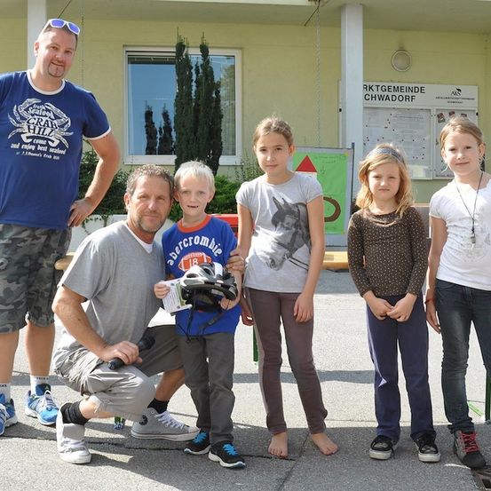 Eine Familie mit fünf Personen, darunter zwei Erwachsene und drei Kinder, steht vor einem Gebäude mit einem Schild, das 'RKTGEMEINDE CHWADORF' besagt. Die Erwachsenen lächeln, und die Kinder posieren für ein Foto. Der Mann auf der linken Seite hält einen Helm.