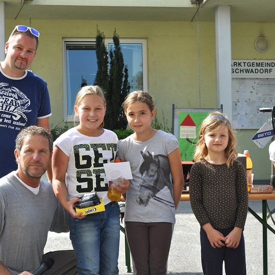 Eine Gruppe von Menschen, darunter zwei Männer und vier Mädchen, steht vor einem Gebäude mit einem Schild, das 'Marktgemeinde Schwadorf' besagt. Der Mann auf der linken Seite hält ein Mikrofon, und das Mädchen auf der linken Seite hält eine gelbe Box mit dem Wort 'Cosy' darauf. Das Mädchen auf der rechten Seite hält eine Flasche.