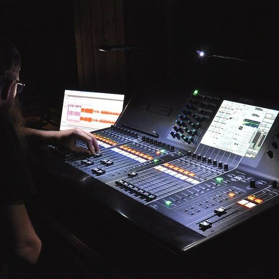 Eine Person justiert ein Soundmischpult mit beleuchteten Tasten und einem Laptop, der eine Audiowelle anzeigt.