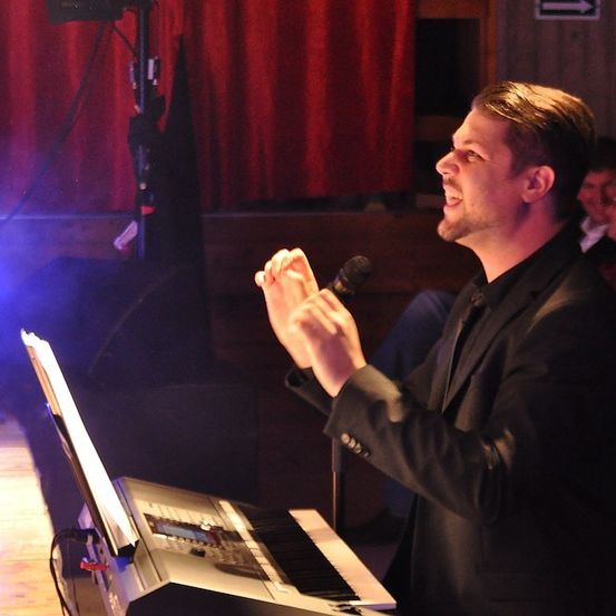 Ein Mann tritt auf der Bühne auf, singt in ein Mikrofon und spielt Klavier. Ein Scheinwerfer beleuchtet ihn, und im Hintergrund sitzen Menschen.