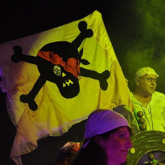 Eine Person in einem Piratenkostüm, die eine Flagge mit Totenkopf und Knochen hält, neben einer anderen Person, die eine weiße Kappe und eine Brille trägt.