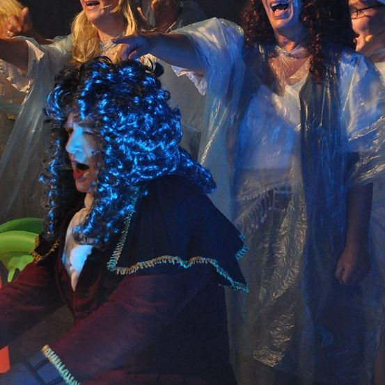 Eine Gruppe von Darstellern auf der Bühne, einer mit blauer Perücke und einer mit lockigem Haar, beide in weißen Kostümen, mit einem Scheinwerferlicht auf sie gerichtet.