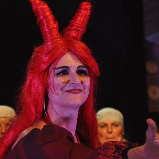 Eine Frau mit roten Haaren und Hörnern auf dem Kopf lächelt und posiert für ein Foto. Sie trägt ein rotes Kleid und scheint auf einer Party zu sein. Hinter ihr tragen zwei andere Frauen weiße Hüte.