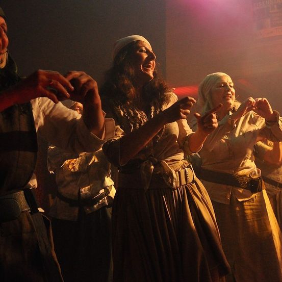 Eine Gruppe von Menschen in traditioneller Kleidung tanzt und lächelt in einem schwach beleuchteten Raum mit lila Beleuchtung.