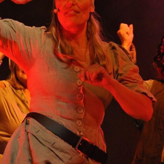 Eine Frau in einem roten Kleid und einem weißen Stirnband tritt auf der Bühne auf, macht Handgesten und lächelt. Sie trägt einen schwarzen Gürtel mit einer Schnalle. Hinter ihr treten auch zwei andere Personen auf.