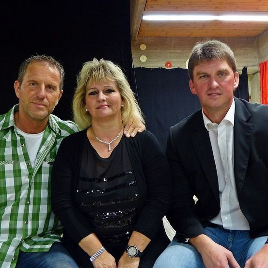 Drei Menschen posieren für ein Foto. Die Frau trägt eine Halskette und eine Uhr. Der Mann links trägt ein grün-weiß kariertes Hemd. Der Mann rechts trägt einen schwarzen Anzug.