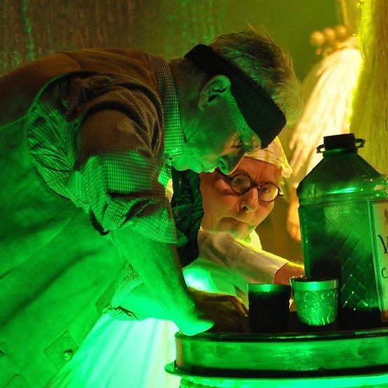 Zwei Männer in einem grün beleuchteten Raum, einer mit einer Augenbinde, gießen eine grüne Flüssigkeit aus einer Flasche in einen Becher.