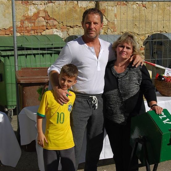 Ein Mann, eine Frau und ein Junge stehen gemeinsam vor einer Ziegelmauer mit einer grünen Box und einem Korb mit Gemüse. Der Junge trägt ein gelbes Fußballtrikot mit der Nummer 10.