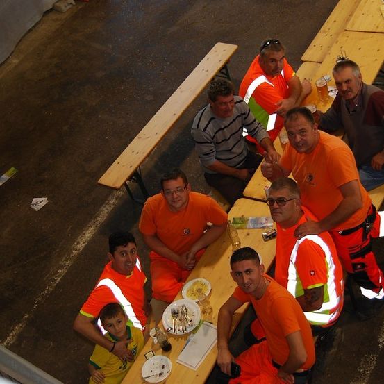 Eine Gruppe von Männern in orangefarbenen Uniformen sitzt auf Bänken in einem Raum, einige trinken und andere essen. Es gibt Teller, Gläser und andere Gegenstände auf den Bänken.