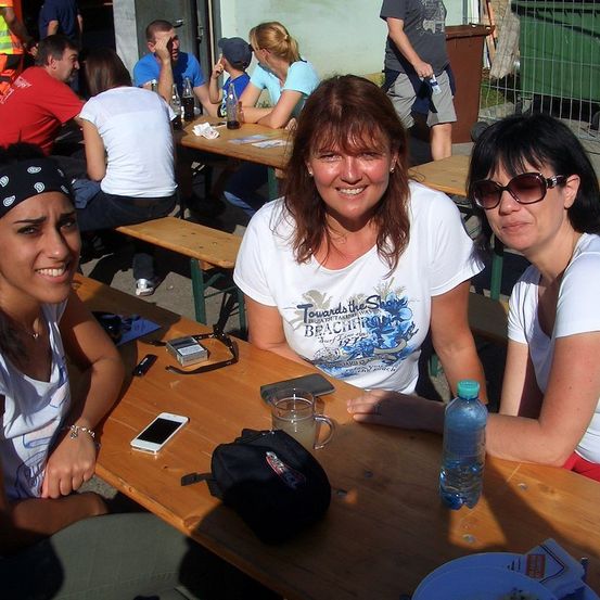 Drei Frauen sitzen an einem Holztisch und lächeln für ein Foto. Eine hat eine Bandana, eine andere trägt eine Sonnenbrille. Eine Wasserflasche, ein Handy und ein Becher stehen auf dem Tisch. Dahinter sitzen weitere Personen an Tischen, einige mit Getränken und Essen.