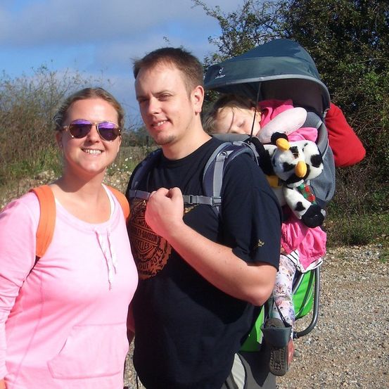Ein Mann und eine Frau stehen neben einem Kinderwagen mit einem schlafenden Kind und einem Plüschtier. Der Mann trägt einen Rucksack und ein Harley Davidson-Shirt.