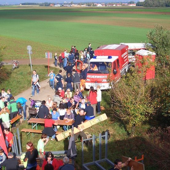 Eine Menschenmenge versammelt sich um einen Feuerwehrwagen und mobile Toiletten auf einem Rasenfeld mit einem großen grünen Feld im Hintergrund.