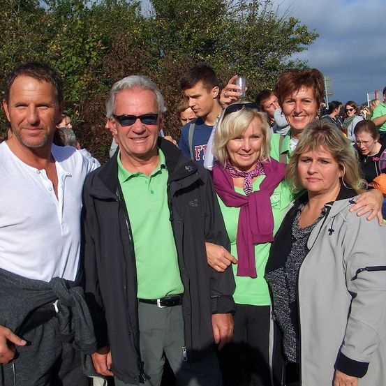 Eine Gruppe von Menschen, darunter zwei Männer und zwei Frauen, stehen zusammen im Freien, lächeln und posieren für ein Foto. Sie sind von einer Menge Menschen umgeben, und im Hintergrund sind Bäume und ein bewölkter Himmel zu sehen.
