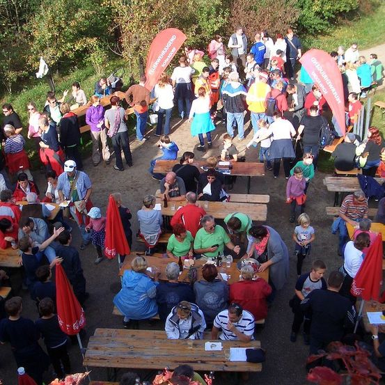 Eine große Gruppe von Menschen versammelt sich bei einer Outdoor-Veranstaltung, sitzt um Holztische unter roten Sonnenschirmen. Einige essen, während andere stehen und reden. Die Umgebung scheint ein Park oder ein Outdoor-Festival zu sein.
