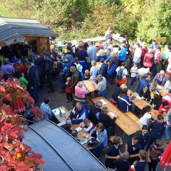 Eine große Menschenmenge versammelt sich um Holztische in einer Freiluftumgebung, möglicherweise bei einem Festival. Einige sitzen, andere stehen, und es gibt Bäume und ein kleines Gebäude im Hintergrund.