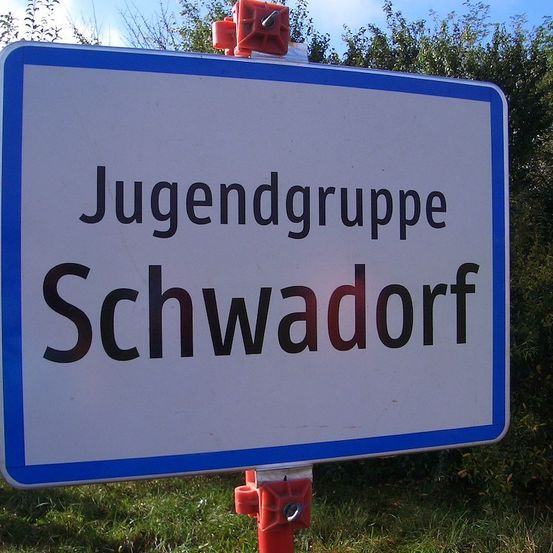 Ein weißes Schild mit blauen Rändern trägt in fetten schwarzen Buchstaben Jugendgruppe Schwadorf.