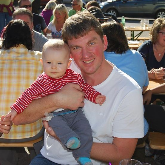 Ein Mann hält ein Baby in einem Restaurant und lächelt, während andere Leute an Tischen im Hintergrund sitzen.