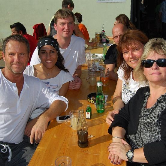 Eine Gruppe von Menschen sitzt an einem Tisch in einem Restaurant, lächelt und posiert für ein Foto. Auf dem Tisch stehen Flaschen, Gläser und andere Gegenstände.