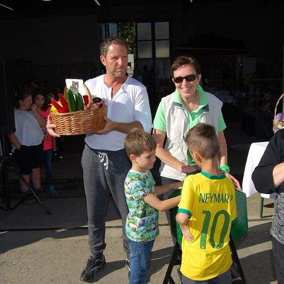 Ein Mann mit einem Korb voller Gemüse steht neben zwei Jungen, von denen einer ein Neymar-Trikot trägt. Eine Frau in einer grünen Weste lächelt hinter ihnen. Im Hintergrund sind Kinder und Erwachsene anwesend.