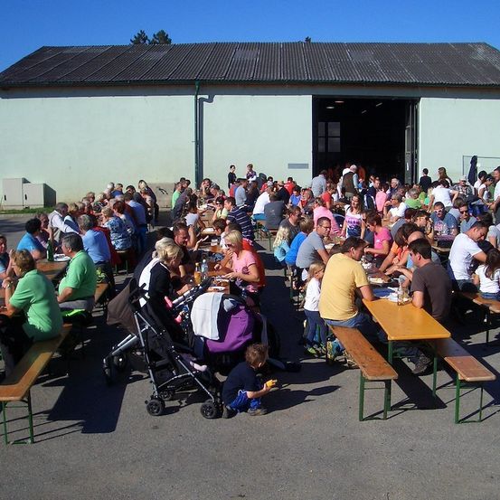 Eine große Gruppe von Menschen sitzt an Picknicktischen vor einem Gebäude, einige essen und andere unterhalten sich. Die Tische sind in Reihen angeordnet, und das Gebäude hat eine große offene Tür.