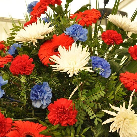 Eine farbenfrohe Blumenarrangement mit roten, weißen und blauen Blumen, einschließlich Gänseblümchen, Nelken und Kornblumen, vor einem unscharfen Hintergrund.